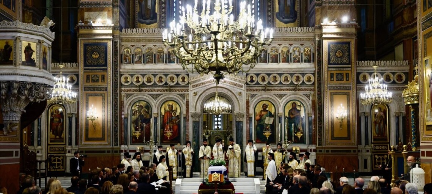 A Greek Orthodox funeral service before the iconostasis, with clergy and a casket